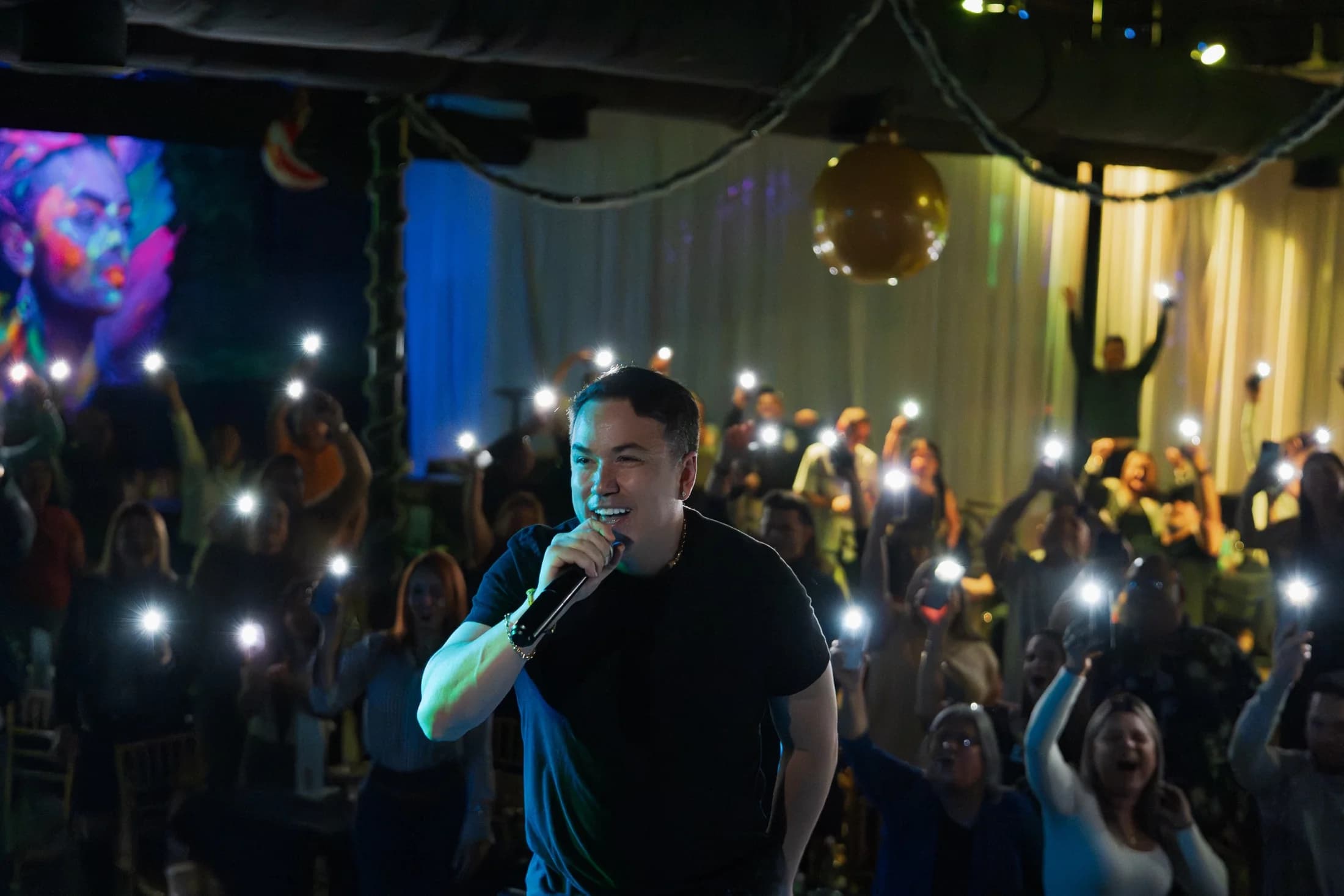 A microphone performer facing an audience holding illuminated phones in a dark room