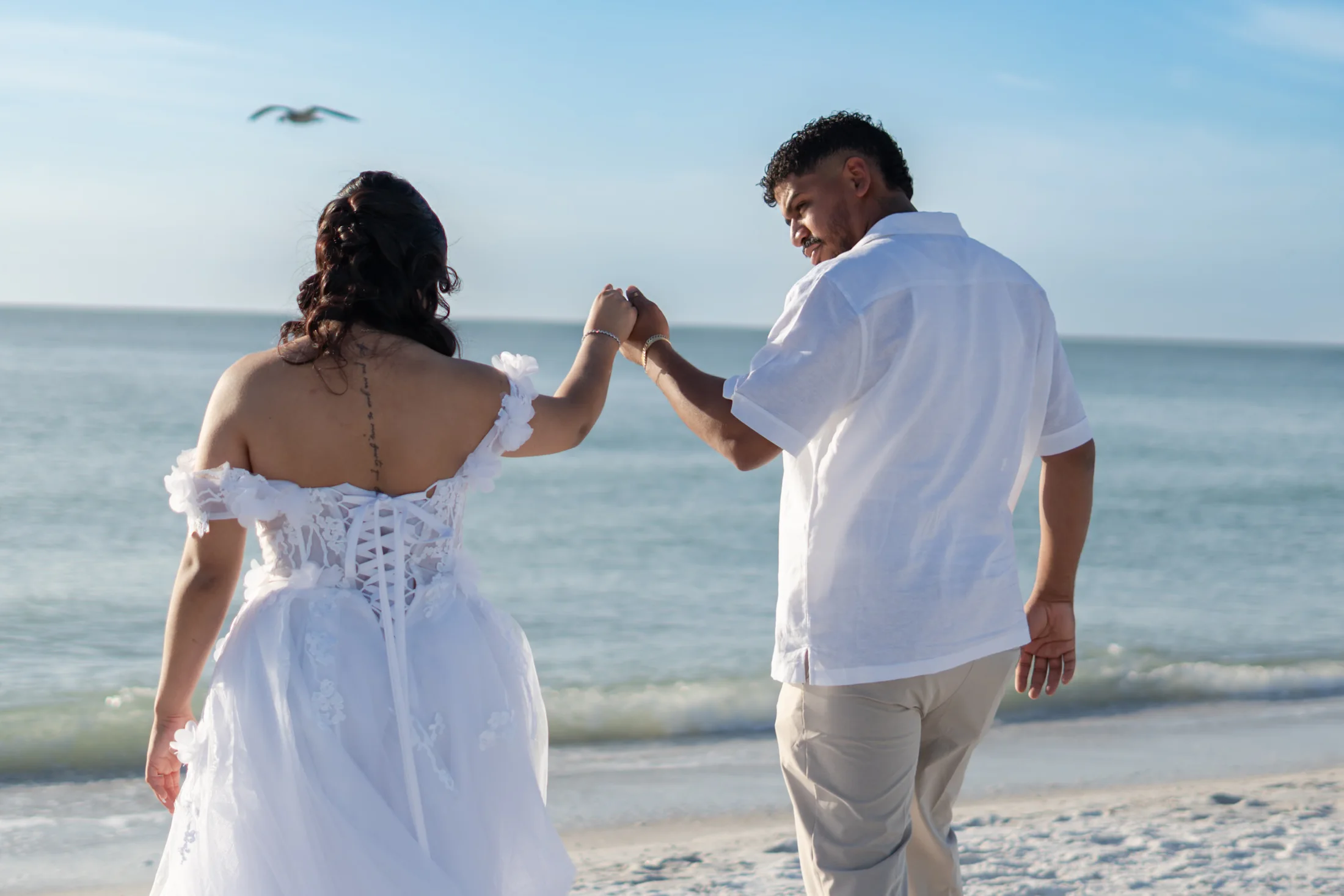 Two people in white holding hands by the ocean beneath an open sky